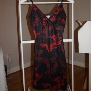 Svart klänning med röda drakar - Supersnygg svart kortklänning med röda drakmönster och smala axelband. Klänningen har omlott-v-ringning och ärmlös design, perfekt för dig som vill sticka ut med en cool och unik stil.