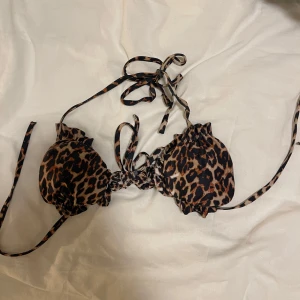 Leopardmönstrad bikini med snörning XS - Snygg bikiniöverdel i leopardmönster med bruna och svarta toner. Triangelformade kupor med volangkant och smala knytband i nacken och ryggen. Tillverkad i stretchigt material med polyester och elastan för perfekt passform. Passar även en S  (den har en fläck på sig se bild) 🫟 