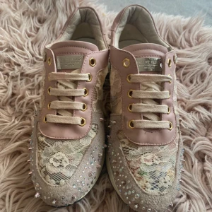 Philipp Plein sneakers med spets och strass - Unika sneakers från Philipp Plein i ljusrosa och beige med spetsdetaljer, blommigt mönster och glittrande strass på sidorna. Skorna har snörning, guldfärgade öljetter och coola nitar. Perfekt för dig som vill sticka ut med en lyxig och feminin stil.