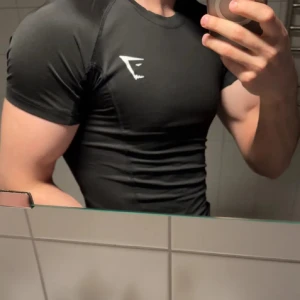 Svart tight t-shirt från Gymshark - Svart t-shirt från Gymshark med vit logga på bröstet. Tight passform som framhäver musklerna, perfekt för gymmet. med korta ärmar och rund halsringning.