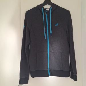 Mörkgrå hoodie från Babolat med blå detaljer - Säljer en mörkgrå hoodie från Babolat med blå dragkedja och snören. Tröjan har huva, långa ärmar och två fickor framtill. Tillverkad i bomull och polyester, perfekt för tennis eller chill dagar. Snygg sportig look med Babolat-logga på bröstet och ärmen.