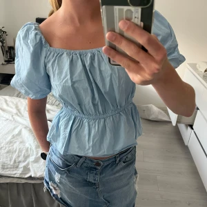 Ljusblå blus med puffärm - Supersöt ljusblå blus med puffiga korta ärmar och smock i midjan som ger en fin volang. Fyrkantig ringning och luftig bomullskänsla, perfekt för varma dagar. Enkel att styla med jeans eller kjol för en fräsch look.