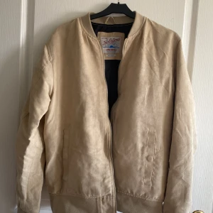 Beige mocka bomberjacka Jack & Jones - Snygg beige bomberjacka från Jack & Jones Originals i storlek medium. Jackan är gjord i mjuk mocka-look, har dragkedja framtill och två fickor. Perfekt för dig som gillar stilren och clean streetwear.