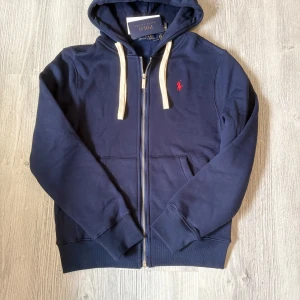 Marinblå hoodie från Polo Ralph Lauren - Snygg marinblå hoodie från Polo Ralph Lauren med dragkedja, vit snörning i huvan och röd broderad logga på bröstet. Mjuk insida och klassisk passform, perfekt för chill dagar. Stora fickor framtill och ribbade muddar för extra komfort. Den har en QR-kod och prislappen kvar som bevisar att den är äkta.