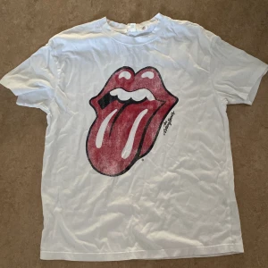 Rolling Stones vit t-shirt med tryck - Vit t-shirt med det ikoniska Rolling Stones-trycket framtill, stora röda läppar och tunga. Klassisk rund hals och korta ärmar. Perfekt för dig som vill ha en cool och tidlös bandtröja med streetkänsla.