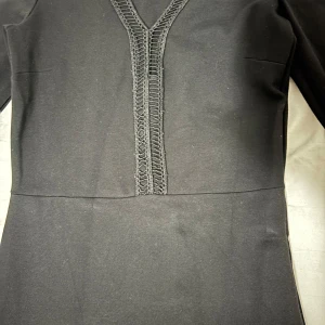 Svart långklänning med spetsdetalj - Stilren svart långklänning med långa ärmar och snygg v-ringning med dekorativ spetsdetalj framtill. Klänningen har en figurnära passform och är perfekt för dig som gillar en klassisk look med en twist. Materialet känns mjukt och följsamt.