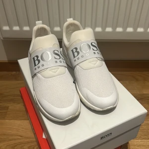 Vita sneakers från Hugo Boss - Snygga vita sneakers från Hugo Boss med bred elastisk rem över foten och stor logga framtill. Ovandelen är i mesh och syntet för en sportig look. Skorna har rund tå, platt sula och dragflik bak för enkel påtagning. Perfekta för dig som gillar stilrena och moderna sneakers. - endast använda en gång, extremt sköna skor. Låda medföljer. Köptes för 2500kr