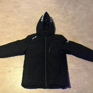 Svart pufferjacka från FSBN CNTMP - Svart pufferjacka med huva från FSBN CNTMP. Jackan har dragkedja framtill, en bröstficka med dragkedja och tryckta vita logodetaljer på huva, axel och rygg. Fodrad insida för extra värme och klassisk pufferlook.