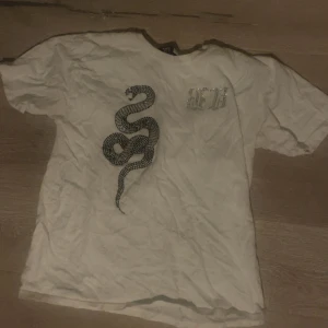 Vit t-shirt med ormmotiv från Bvaldi - Vit t-shirt från Bvaldi i storlek XS med korta ärmar. Framsidan har ett stort ormmotiv i glittrande paljetter och text i silvriga stenar. Skön passform och cool design som sticker ut. Perfekt för dig som gillar edgy detaljer och streetstyle.
