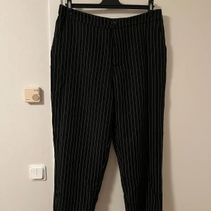 Svarta kostymbyxor med vita ränder - Snygga svarta kostymbyxor med smala vita ränder i klassiskt pinstripe-mönster. Byxorna har normal passform, hällor för bälte och stängs med knapp och dragkedja. Perfekta för en stilren och trendig look.Katt finns i hemmet 