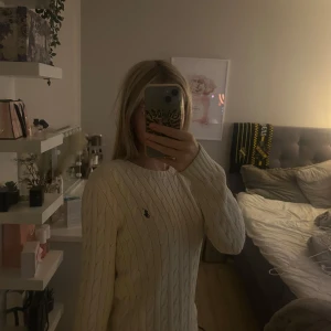 Beige Ralph Lauren Tröja - Kabelstickad beige tröja från Ralph Lauren i 100% bomull. Klassisk rund halsringning och långärmad modell med den ikoniska broderade loggan på bröstet.