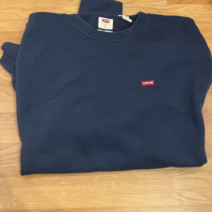 Mörkblå sweatshirt från Levi's - En stilren mörkblå sweatshirt från Levi's med klassisk rund hals och diskret röd logotyp på bröstet. Tröjan har långa ärmar och ribbade muddar vid ärmslut och nederkant. Perfekt för en chill och enkel look.