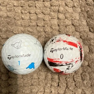 Rare taylormade bollar - Bilden visar två golfbollar från TaylorMade, en med blå och grå handtecken och en med röd och svart design. 