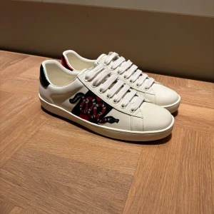 Gucci Ace  - Skorna har nya snören på sig!           Gucci Ace sneakers i vitt skinn och en röd och svart orm på sidan. Baksidan har metallicröd och metallicgrön detalj med Gucci-logga. Klassisk rund tå, breda vita snören och platt sula. Snygg och lyxig statement-sneaker. 