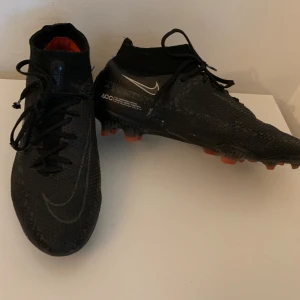 Nike Phantom GT elite fotbollsskor svarta - Svarta Nike Phantom GT fotbollsskor med snörning och högt skaft i syntetmaterial. Skorna har en texturerad ovandel för bättre bollkontroll och orange detaljer på dobbarna. Snygg och modern design med Nike-logga på sidan. Påse kommer ej med.