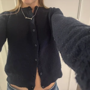 Mörkblå stickad kofta med knappar - Mysig mörkblå stickad kofta med rund hals och knäppning framtill. Koftan har långa ärmar och en lite kortare passform som ger en chill vibe. Perfekt att slänga över en t-shirt eller linne för extra värme.