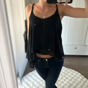 Svart flowy linnetopp med tunna band - Säljer en svart, luftig linnetopp med tunna axelband och snyggt fall. Toppen har dubbla lager i transparent polyester och cool detalj i ryggen med flera smala band. Perfekt för dig som gillar en stilren men edgy look.