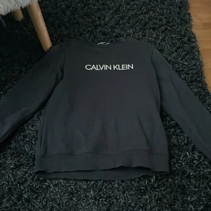 Svart Calvin Klein Jeans sweatshirt - Svart sweatshirt från Calvin Klein Jeans med vit logga framtill och en patch med texten 'JEANS' bak vid nederkanten. Klassisk rund halsringning och långa ärmar. Perfekt för en clean och stilren look. Hör av er vid frågor.