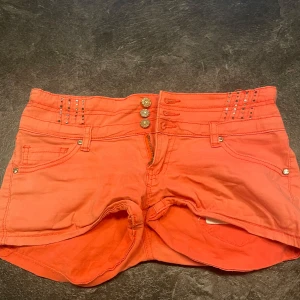 Korallrosa shorts med nitar och knappar - Snygga korallrosa shorts med låg midja, dekorerade med glittriga nitar och tre guldiga knappar framtill. Shortsen har klassiska fickor både fram och bak samt coola sömdetaljer. Perfekta för sommaren! står ingen storlek men skulle säga 34-36!