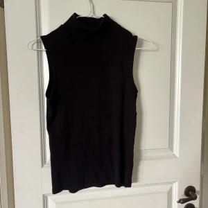 Svart ärmlös polotopp - Snygg svart polotopp utan ärmar med hög krage. Perfekt att styla med jeans eller kjol för en clean look. Toppens material känns mjukt och stretchigt, vilket gör den bekväm att bära hela dagen.