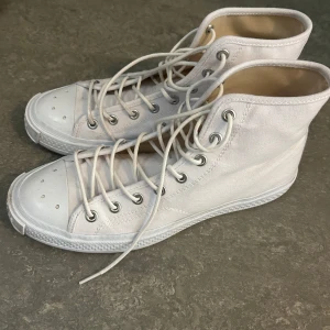 Vita höga sneakers Acne studios - Säljer ett par vita höga sneakers från Acne studios. Skorna har en platt sula och är i canvas med gummidetaljer på tån. Metallöglor och stilren design gör dem perfekta för en clean och fräsch look.