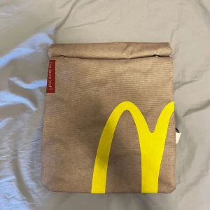 McDonald's väska/ryggsäck - Unik väska i brunt tyg med McDonald's ikoniska gula M-logga och röd etikett med texten 'I'm lovin' eat'. Väskan har rullstängning och knappar för extra säkerhet. Perfekt för dig som vill sticka ut med en cool och annorlunda accessoar. Vid frågor eller liknande är det bara att höra av sig! 