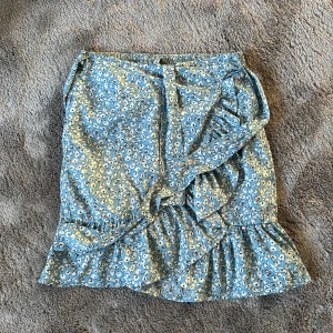 Blommig blå volangkjol från ONLY - Supersöt kort kjol från ONLY i ljusblått med vitt och svart blommönster. Kjolen har volangkant nertill och omlott-design med knyt i midjan. Perfekt för vår och sommar, och den elastiska midjan gör den bekväm att bära hela dagen.