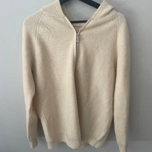 Beige stickad hoodie med half zip - Mysig beige stickad hoodie med half zip och huva. Tröjan har en ribbad struktur och långa ärmar, perfekt för lager-på-lager under kyliga dagar. Enkel och stilren design som funkar till det mesta.