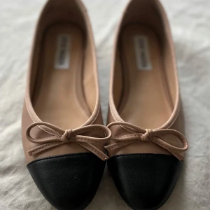 Beige ballerinaskor från Steve Madden - Snygga ballerinaskor från Steve Madden i beige skinn med svart tå och dekorativ rosett framtill. Skorna har rund tå och platt sula, samt en liten metallknapp med logga bak. Perfekta för en chic och klassisk look.