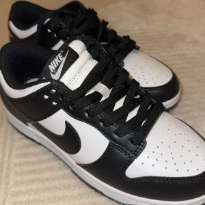 Nike Dunk Low svart/vit sneakers - Snygga Nike Dunk Low sneakers i svart och vitt skinn. Klassisk siluett med svart swoosh, perforerad tå och svart snörning. Perfekta för dig som gillar streetstyle och vill ha en ikonisk look. HELT OANVÄNDA