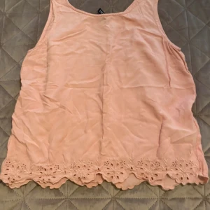 Ljusrosa linnetopp från Divided , S  - Supersöt ljusrosa linnetopp från Divided med broderade spetsdetaljer längs nederkanten. Toppen är ärmlös och har en loose passform, perfekt för varma dagar. Materialet känns lätt och luftigt, och färgen är mjukt rosa.