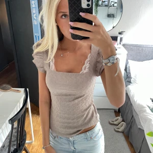 Beige smockad topp med volangärm - Supersöt beige topp med smockat tyg och fyrkantig urringning. Korta volangärmar och spetsdetalj i vitt vid halsen ger en romantisk vibe. Perfekt att styla med jeans eller kjol för en trendig look.