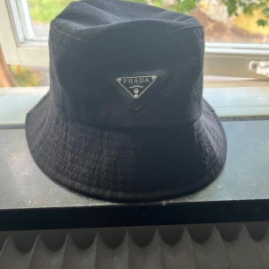 Svart bucket hat från Prada - Stilren svart bucket hat från Prada med klassisk siluett och ikonisk metallogo framtill. Hatten är tillverkad i ett mjukt tyg som ger en avslappnad vibe och passar perfekt till streetwear eller när du vill lyfta din outfit.