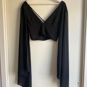 Svart croppad omlottopp med vida ärmar - Säljer en svart croppad omlottopp med långa, vida ärmar och djup v-ringning både fram och bak. Toppen har tunna korsade band i ryggen som ger en snygg detalj. Perfekt för dig som gillar en edgy och trendig stil. Har använts på många festivaler och slår aldrig fel! Har fått en liten defekt som syns på bild, men annars i fint skick!