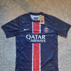 PSG 24/25 Champions League Tröja - Paris Saint-Germain fotbollströja från Nike i marinblått med röd och vit vertikal rand på framsidan. PSG-logga och Nike Swoosh på bröstet, Qatar Airways-tryck och gul stjärna ovanför klubbmärket. Tillverkad i lätt och ventilerande material.