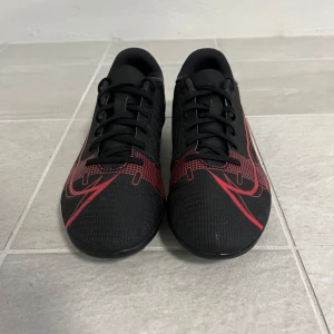 Nike Mercurial svarta fotbollsskor - Svarta Nike Mercurial fotbollsskor med röda detaljer och grafiska mönster. Skorna har snörning, låg profil och dobbar i gult och svart för bra grepp på planen. Snygg sportig look med klassisk Swoosh-logga på sidan. Perfekta för dig som vill sticka ut på fotbollsplanen.