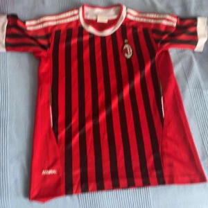 AC Milan Tröja - Snygg fotbollströja från Admiral med klassiska röda och svarta vertikala ränder, vita detaljer vid krage och ärmslut samt tre vita ränder på axlarna. AC Milan-liknande design med klubbmärke på bröstet. Tillverkad i lätt och ventilerande material, perfekt för match eller träning.