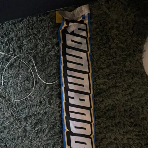 IFK Göteborg halsduk  - Säljer en fet supporterhalsduk för IFK Göteborg med texten 'KOMMANDO BRYGGAN' och klubbens emblem. Halsduken har blåvita ränder, stora svarta bokstäver med guldkant och vita fransar i ändarna. Perfekt för dig som vill visa ditt stöd på läktaren.