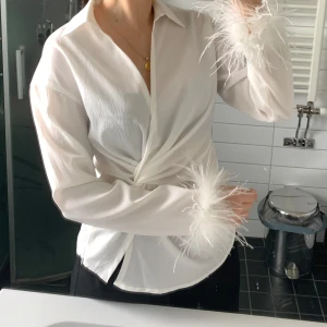 Vit omlottblus med fjädrar - Säljer en vit omlottblus med långa ärmar och fluffiga fjäderdetaljer vid ärmsluten. Helt ny och oanvänd. Passar även S. 