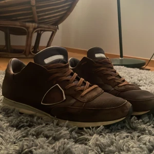 Bruna sneakers från Philippe Model - Snygga bruna sneakers från Philippe Model med klassisk låg siluett och vita detaljer på sidan. Skorna har snörning, platt sula och är tillverkade i en mix av mocka och skinn. Perfekta för dig som gillar en stilren men ändå sportig look.