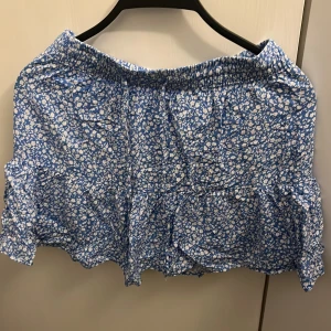 Blommig blå volangkjol från pigalle  - Supersöt kort kjol från AALI i blått med vitt blommönster. Kjolen har volanger och resår i midjan för skön passform. Perfekt för dig som gillar en feminin och lekfull stil. Materialet känns lätt och luftigt, perfekt för varmare dagar.