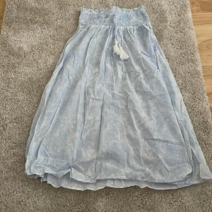 Ljusblå långkjol från Gina Tricot - Supersöt ljusblå långkjol från Gina Tricot med diskret paisleymönster och smock i midjan. Kjolen har knytband med tofsar framtill och är gjord i ett lätt, flowy material som passar perfekt för varma dagar.