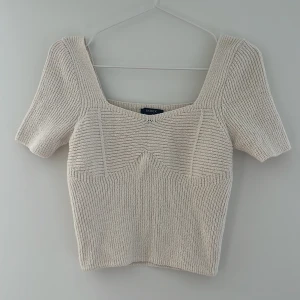 Beige stickad topp från Lindex - Superfin beige stickad topp från Lindex med fyrkantig ringning och korta ärmar. Toppen har en ribbad struktur och markerade sömmar över bysten som ger en snygg form. Perfekt att styla med jeans eller kjol för en trendig look.