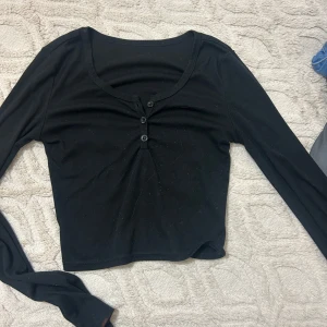 Svart croppad långärmad topp - En svart croppad topp med lång ärm och tre knappar framtill. Djup rundad ringning och enkel, stilren design som passar till det mesta. Mjuk och stretchig känsla, perfekt för lager-på-lager eller som den är.