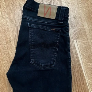 Svarta jeans från Nudie Jeans - Snygga svarta jeans från Nudie Jeans med klassisk femficksdesign och diskret logga på bakfickan. Jeansen har smal passform och är tillverkade i mjukt denimtyg som sitter riktigt bra. Perfekta för dig som gillar stilrena och tidlösa jeans.