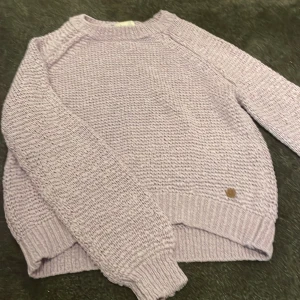 Ljuslila stickad tröja från Ü - Supermysig ljuslila stickad tröja från Ü i storlek 158/164. Tröjan har grovstickad struktur, rund hals och ribbade muddar vid ärmslut och nederkant. Perfekt för lager-på-lager och chill dagar.