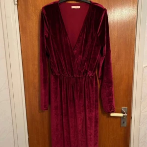 Vinröd sammetsklänning SÄLJES! - Långklänning i vinröd sammet med djup v-ringning och långa ärmar. Klänningen har markerad midja och ett mjukt, glansigt tyg som ger en lyxig känsla. Öppning i bena. Använd en gång. Storlek 36
