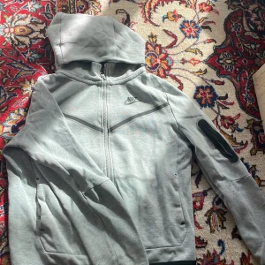 Grå Nike hoodie med dragkedja L - Snygg grå hoodie från Nike i storlek L med hel dragkedja och huva. Tröjan har svarta detaljer vid ärmslut och en praktisk ficka med dragkedja på ena ärmen. Perfekt för chill eller träning, klassisk Nike-logga på bröstet.