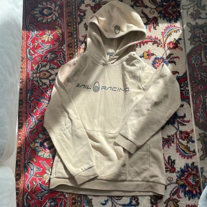Beige hoodie från Sail Racing - Snygg beige hoodie från Sail Racing med svart logga på bröstet och en mindre logga på huvan. Tröjan har känguruficka framtill och är tillverkad i mjuk bomullsmix. Perfekt för chill dagar eller när du vill ha en clean och stilren look.