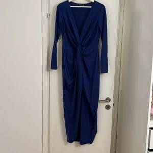 Marinblå långklänning med v-ringning - Snygg marinblå långklänning med djup v-ringning och twistad detalj framtill. Klänningen har långa ärmar och ett elegant fall med drapering över midjan. Perfekt för dig som vill sticka ut med en stilren och modern look.